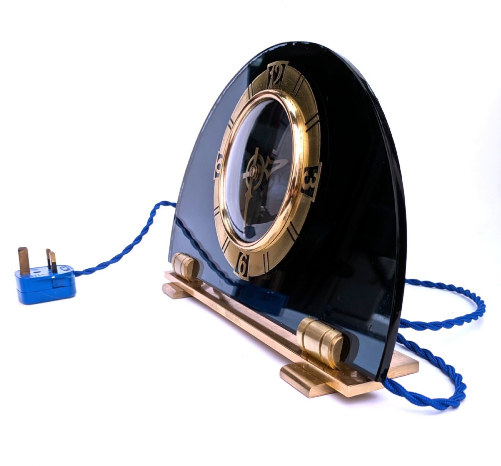 An Art Deco mantel clock featuring a semi-circular blue glass body, a circular brass dial with stylized numerals, and a blue braided flex. View III