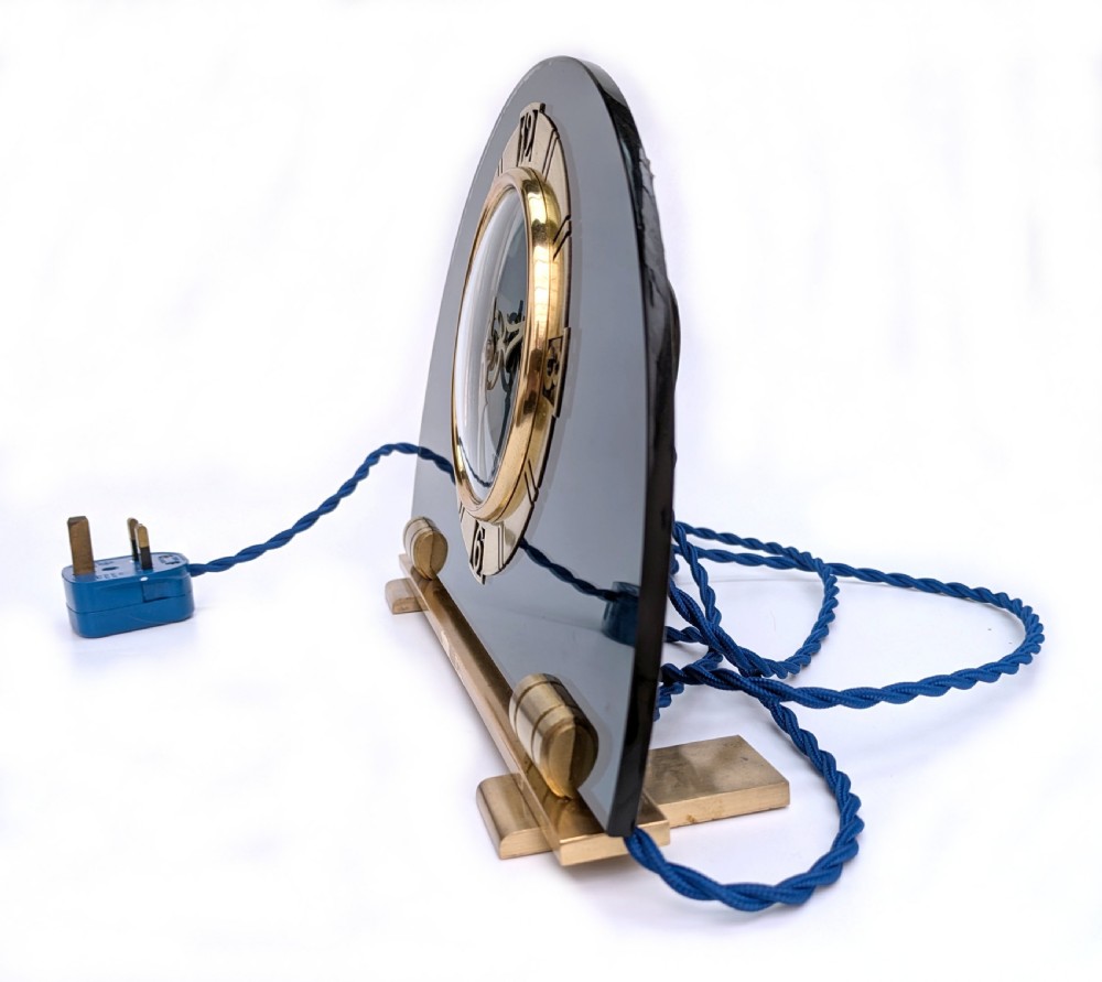 An Art Deco mantel clock featuring a semi-circular blue glass body, a circular brass dial with stylized numerals, and a blue braided flex. View IV
