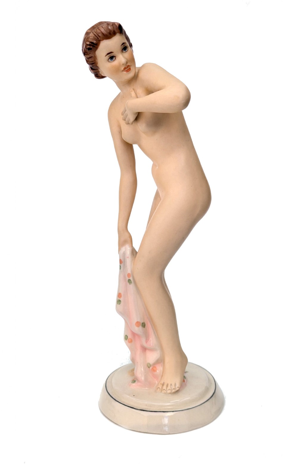 An Art Deco glazed porcelain figurine depicting a standing nude female figure holding a soft pink patterned drape on a rounded cream base.