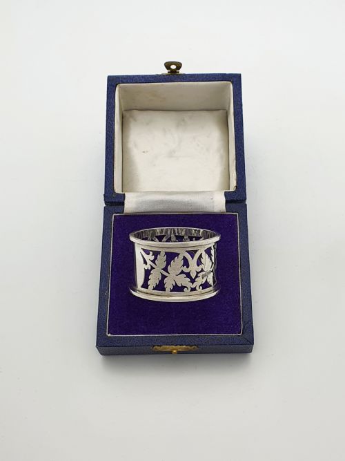 fig 1. An Edwardian sterling silver napkin ring featuring intricate pierced leaf motifs and an unengraved central oval cartouche, presented in a velvet-lined blue box.