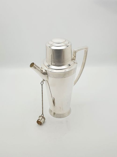 fig 1. An Art Deco silver-plated cocktail shaker featuring a tapered body with concentric ring engravings, a spout with a chained cap, and an angular handle.