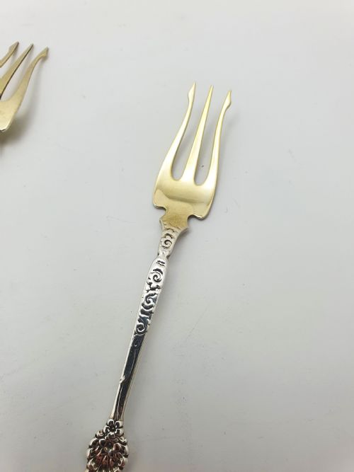 fig 1. A set of twelve sterling silver seafood forks with ornate floral handles and gold gilt tines arranged in a diagonal line.