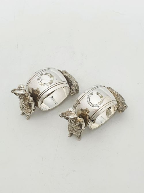 fig 1. A pair of Victorian silver-plated napkin rings featuring detailed cast figures of young girls wearing bonnets standing beside engraved circular rings.