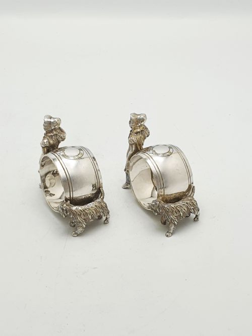 fig 2. A pair of Victorian silver-plated napkin rings featuring detailed cast figures of young girls wearing bonnets standing beside engraved circular rings.