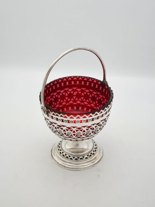 fig 1. An Edwardian silver-plated footed basket featuring pierced geometric fretwork, a swing handle, and a removable ruby red glass liner.