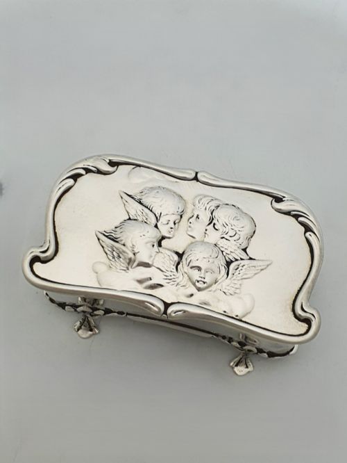 fig 1. An Edwardian sterling silver trinket box featuring an embossed lid depicting cherub heads and a scrolling border, supported by four cast feet.