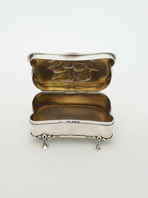 fig 2. An Edwardian sterling silver trinket box featuring an embossed lid depicting cherub heads and a scrolling border, supported by four cast feet.