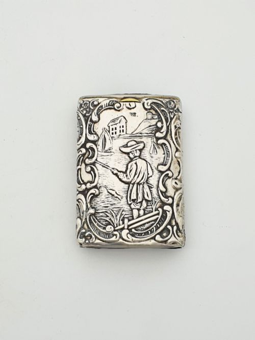 fig 1. A rectangular silver vesta case with detailed embossed scrollwork around a scenic relief of a figure by water, featuring a sliding gilt interior compartment.