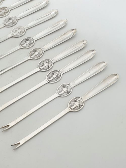 fig 1. A set of twelve silver-plated lobster picks featuring embossed lobster medallions on the handles and narrow two-pronged tips, arranged neatly in a diagonal row.