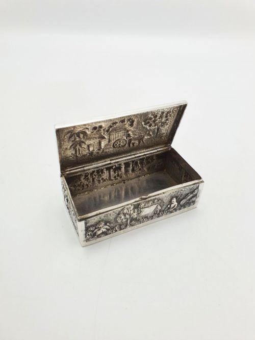 fig 1. A rectangular antique silver trinket box featuring detailed repousse relief depicting a village scene with huts and palm trees.