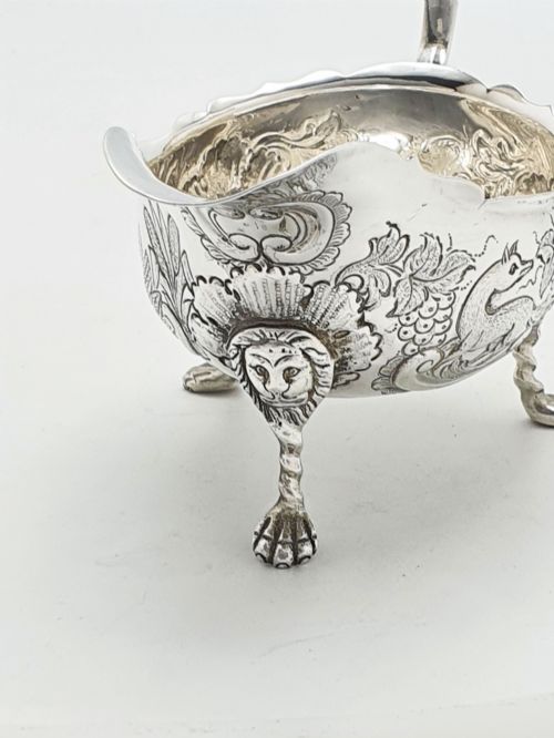 fig 1. A pair of engraved silver-plated sauce boats with curved handles and decorated cabriole legs resting on a neutral background.
