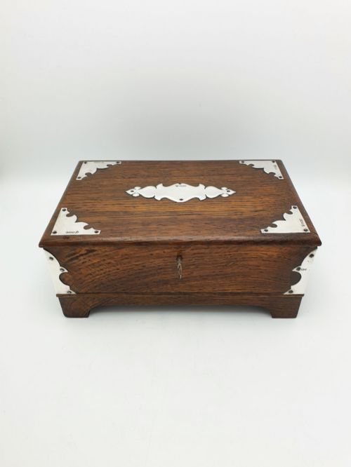 fig 2. An Edwardian oak cigar box featuring decorative sterling silver corner mounts, a central shaped silver plaque, and a front keyhole with key.