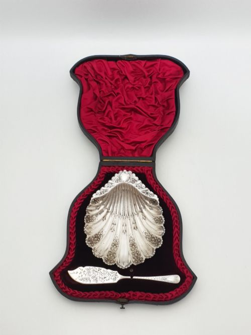 fig 1. A Victorian sterling silver scallop shell butter dish and matching engraved spreader knife resting inside a fitted red and black velvet presentation case.