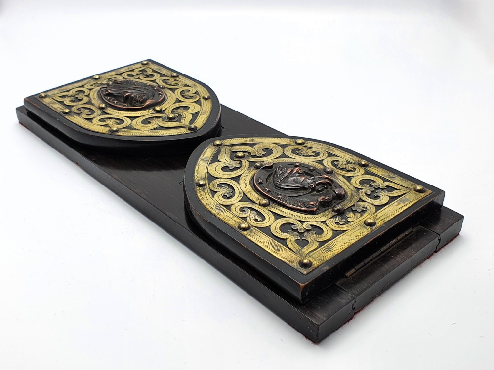 An ebonised wooden expanding book slide featuring pointed arched folding ends decorated with engraved brass straps and a central copper horseshoe medallion with a horse profile. View II