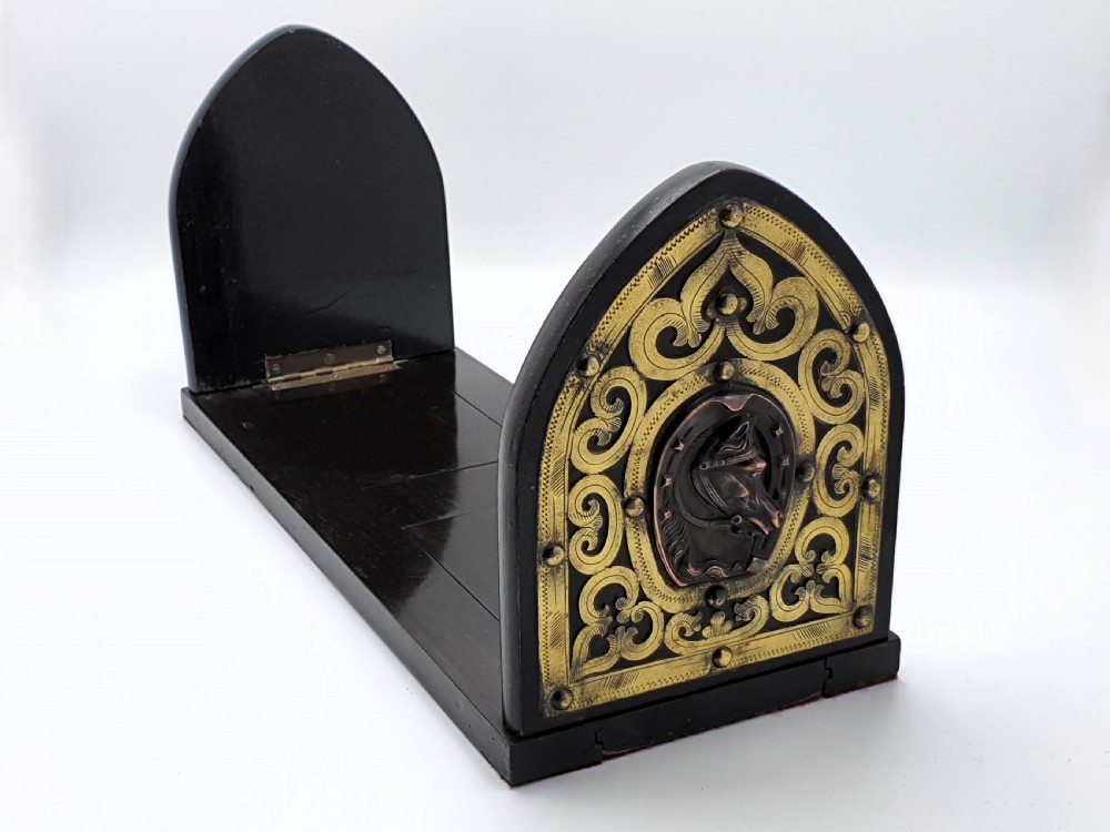 An ebonised wooden expanding book slide featuring pointed arched folding ends decorated with engraved brass straps and a central copper horseshoe medallion with a horse profile. View III