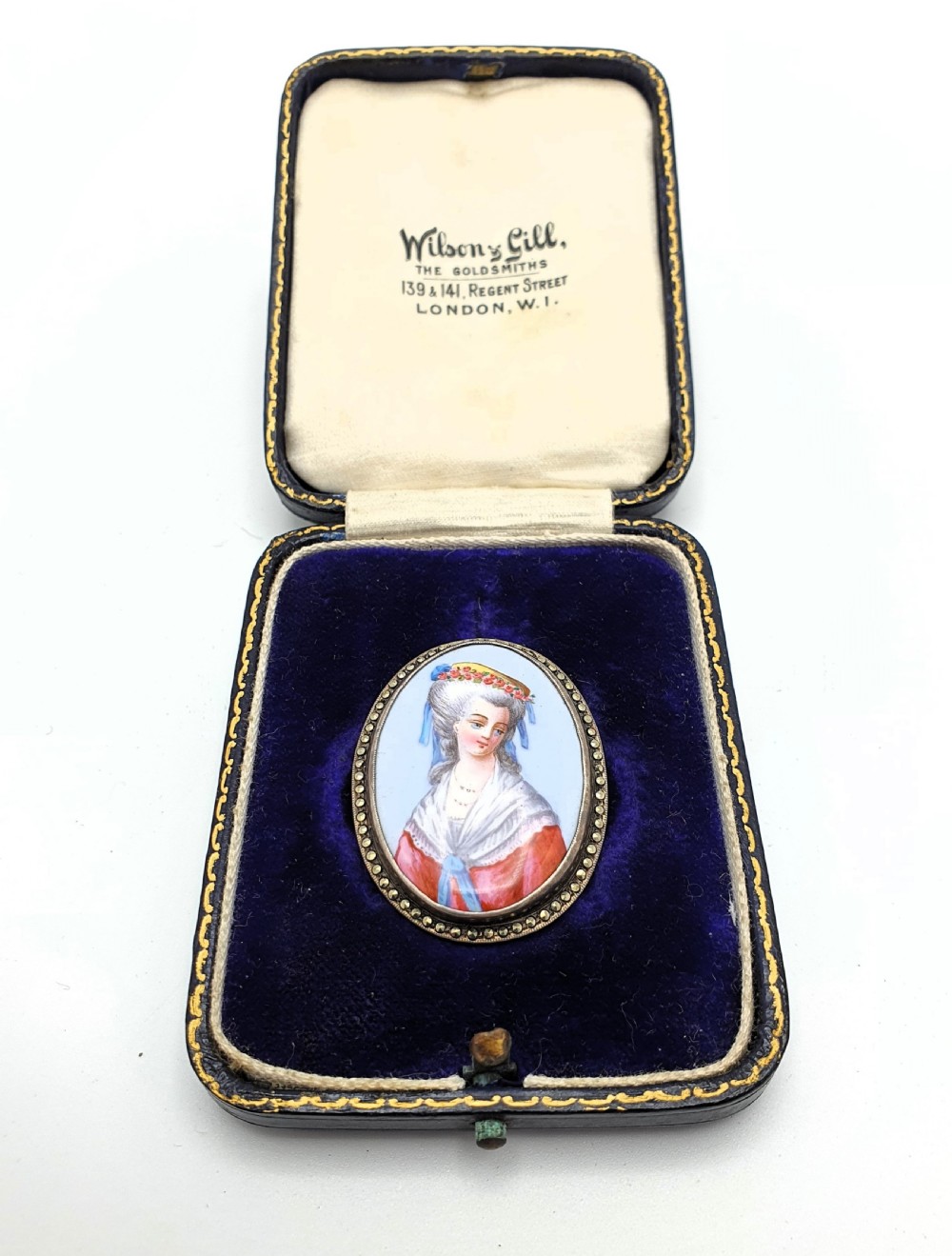An oval silver pendant brooch featuring a hand-painted portrait of an eighteenth-century woman, presented inside a blue velvet box.