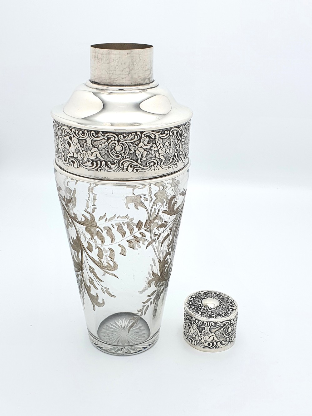An antique clear glass cocktail shaker featuring etched silver overlay foliage patterns and relief-carved sterling silver mounts depicting cherubs. View VI