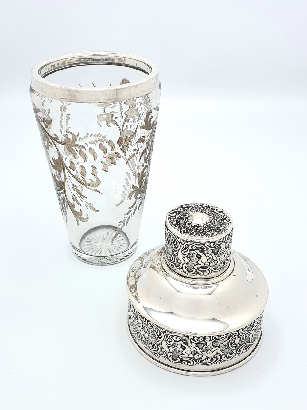 An antique clear glass cocktail shaker featuring etched silver overlay foliage patterns and relief-carved sterling silver mounts depicting cherubs. View VII