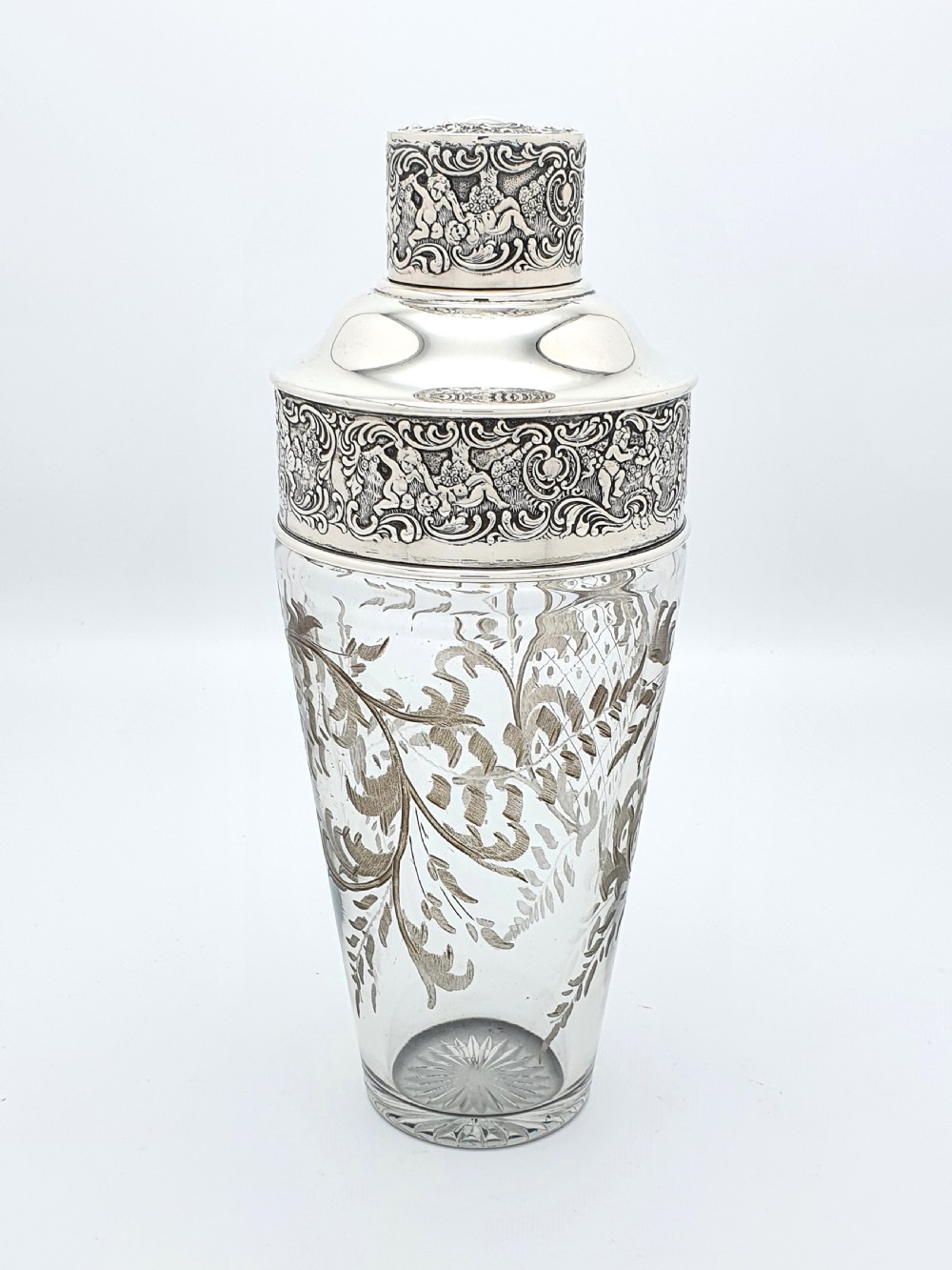 An antique clear glass cocktail shaker featuring etched silver overlay foliage patterns and relief-carved sterling silver mounts depicting cherubs.