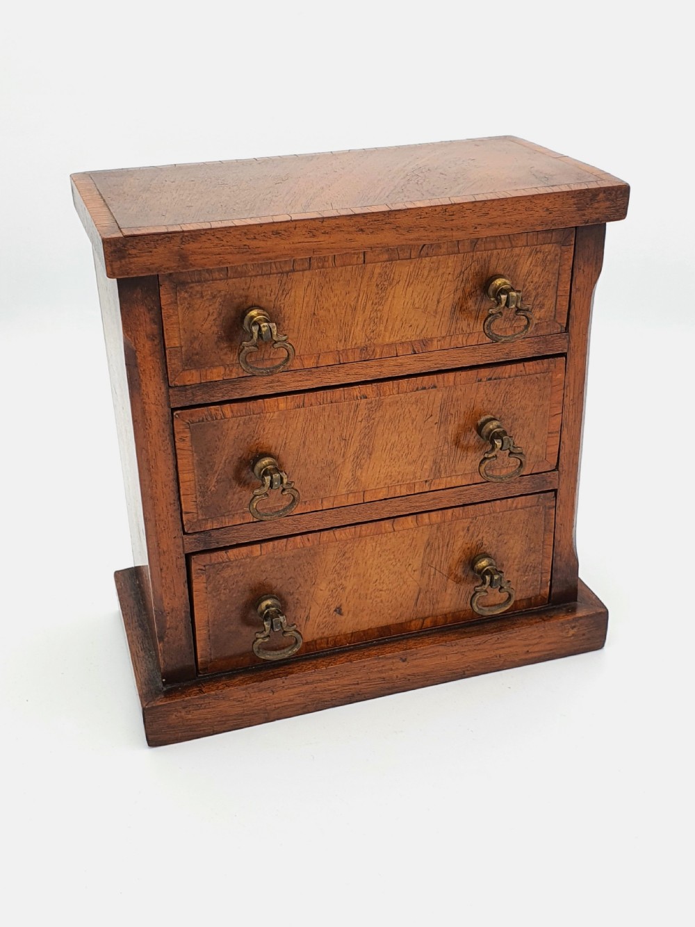 A small mahogany three-drawer miniature chest with brass drop handles and inlaid wood banding.