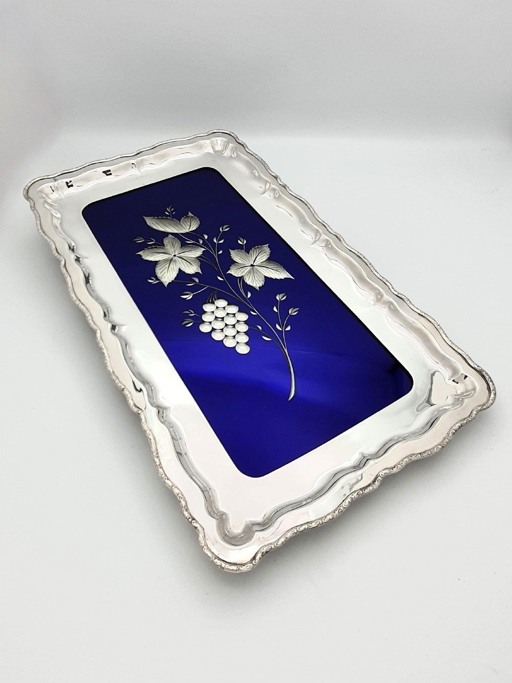 A rectangular silver-plated tray with a scalloped border featuring a deep cobalt blue glass mirror insert engraved with a grapevine and leaf motif.