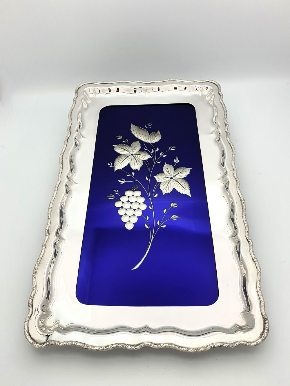 A rectangular silver-plated tray with a scalloped border featuring a deep cobalt blue glass mirror insert engraved with a grapevine and leaf motif. View II
