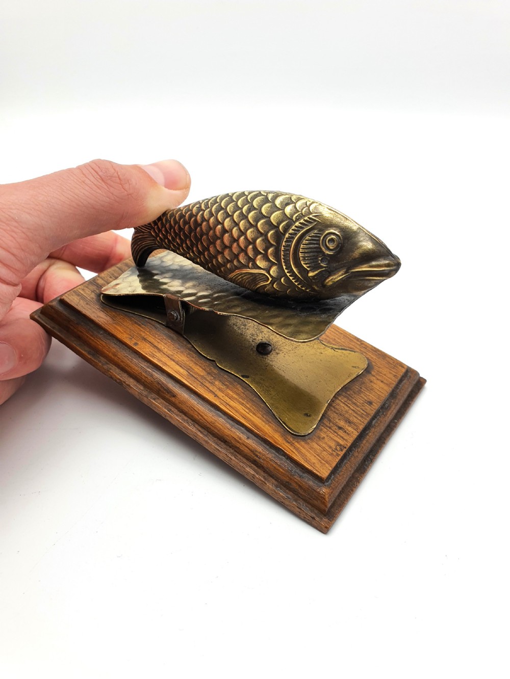 A 1920s brass fish-shaped spring clip mounted onto a molded rectangular wooden base.