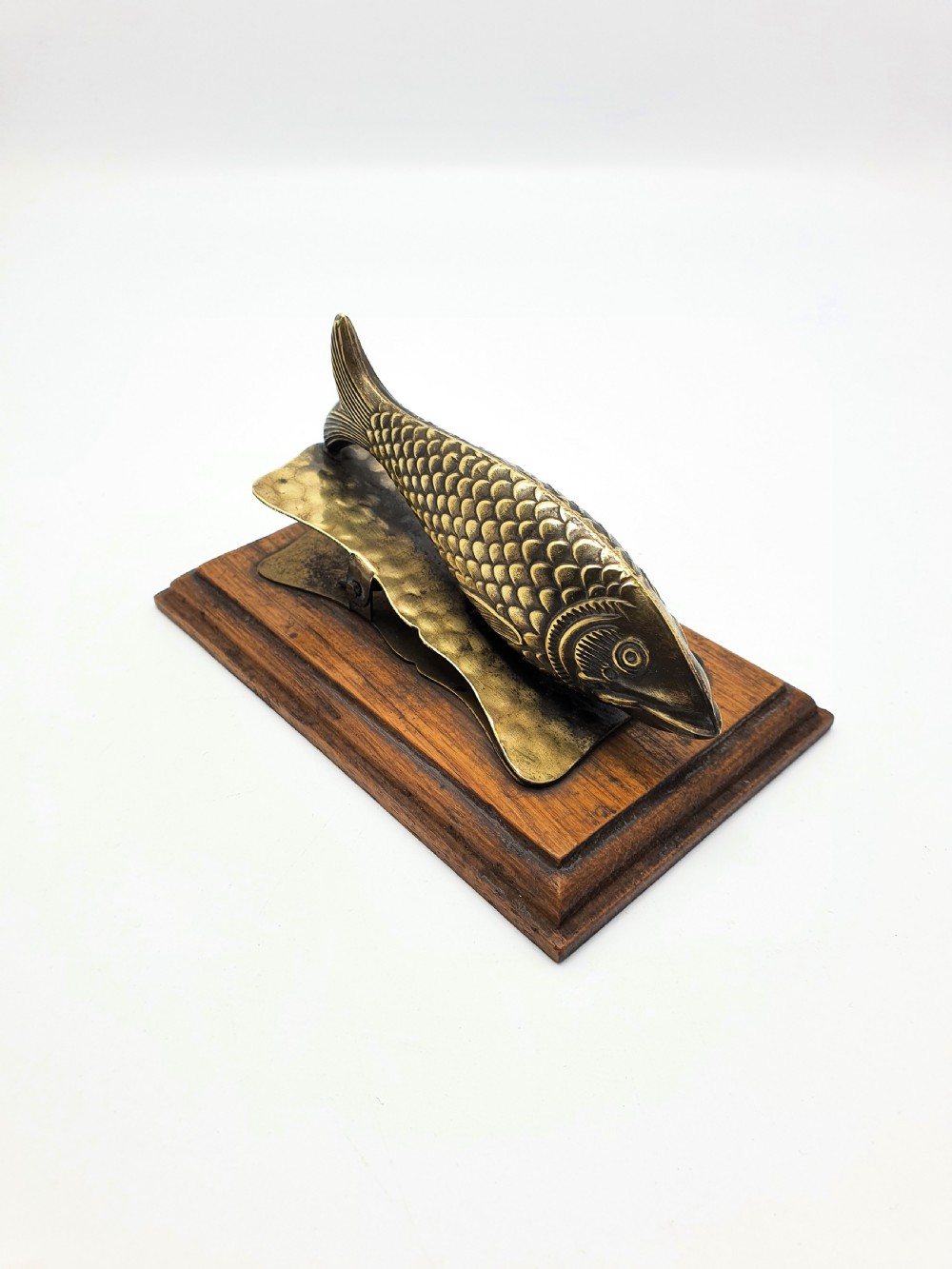 A 1920s brass fish-shaped spring clip mounted onto a molded rectangular wooden base. View II