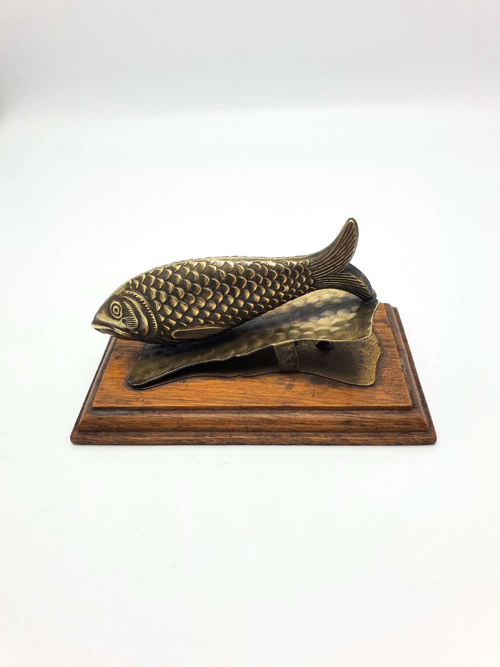 A 1920s brass fish-shaped spring clip mounted onto a molded rectangular wooden base. View III