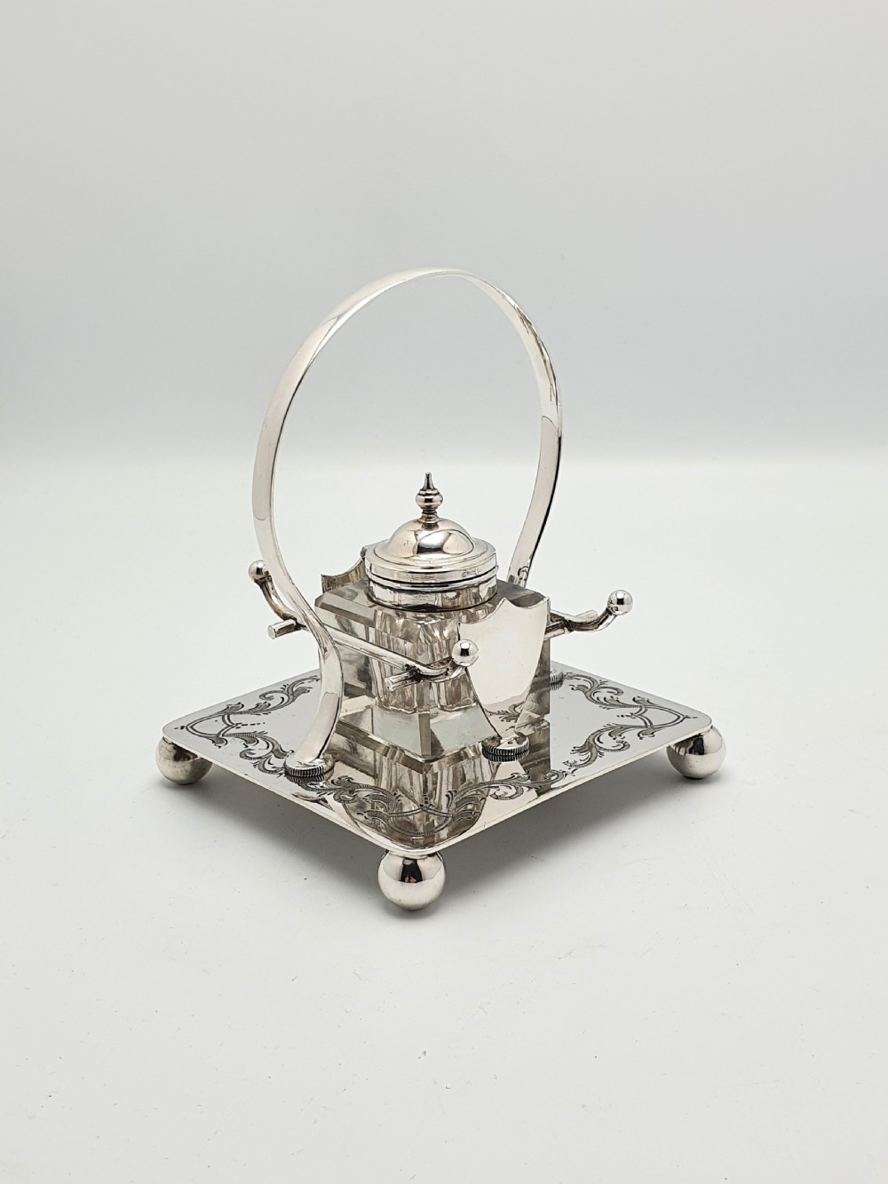A Victorian silver-plated inkstand featuring an engraved square base on ball feet, a central clear cut-glass inkwell with a silver lid, an overhead hoop handle, and side pen rests.
