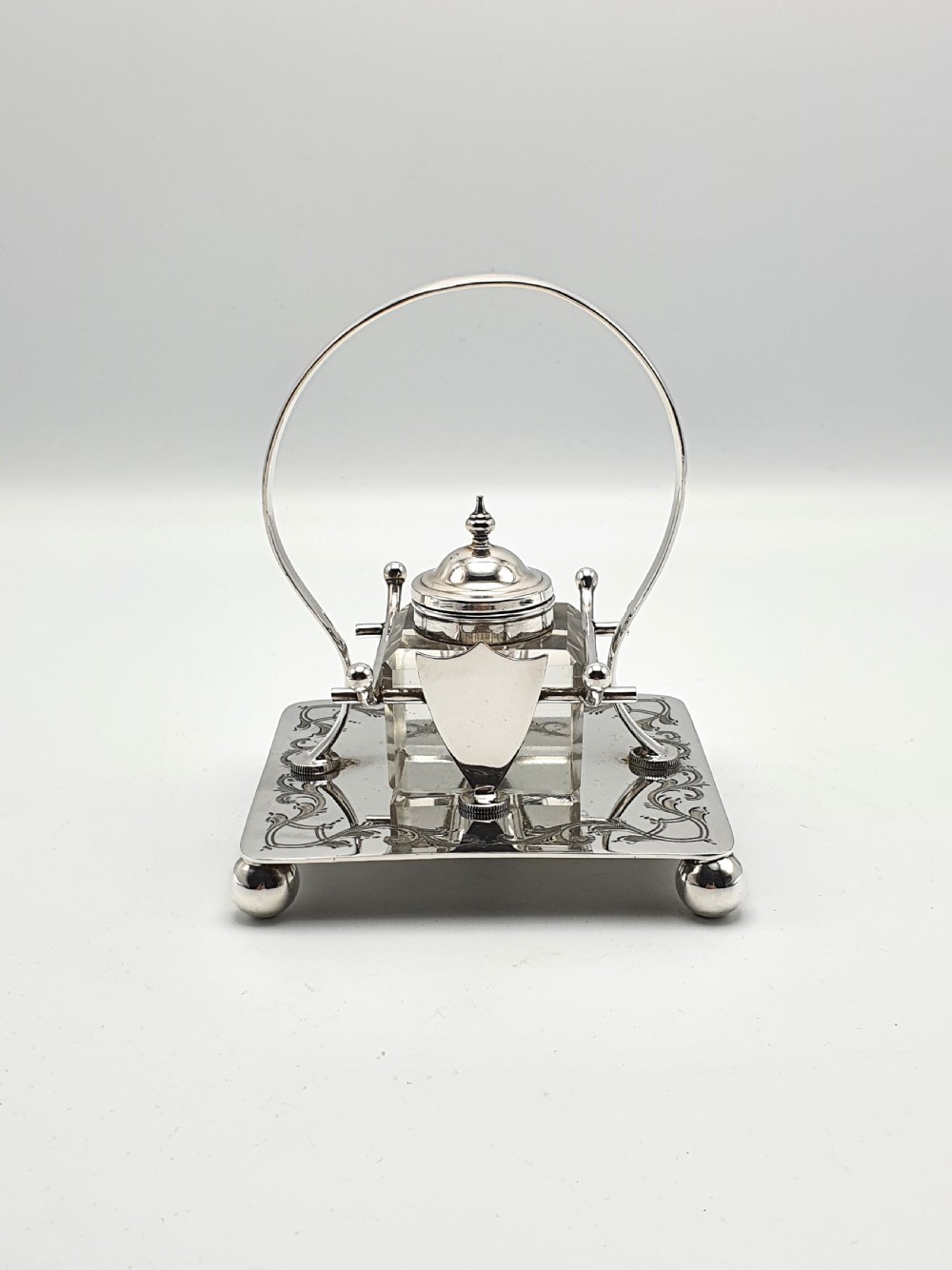 A Victorian silver-plated inkstand featuring an engraved square base on ball feet, a central clear cut-glass inkwell with a silver lid, an overhead hoop handle, and side pen rests. View II