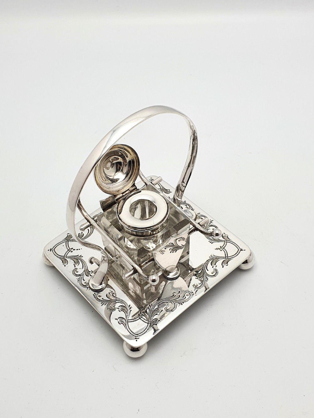 A Victorian silver-plated inkstand featuring an engraved square base on ball feet, a central clear cut-glass inkwell with a silver lid, an overhead hoop handle, and side pen rests. View III