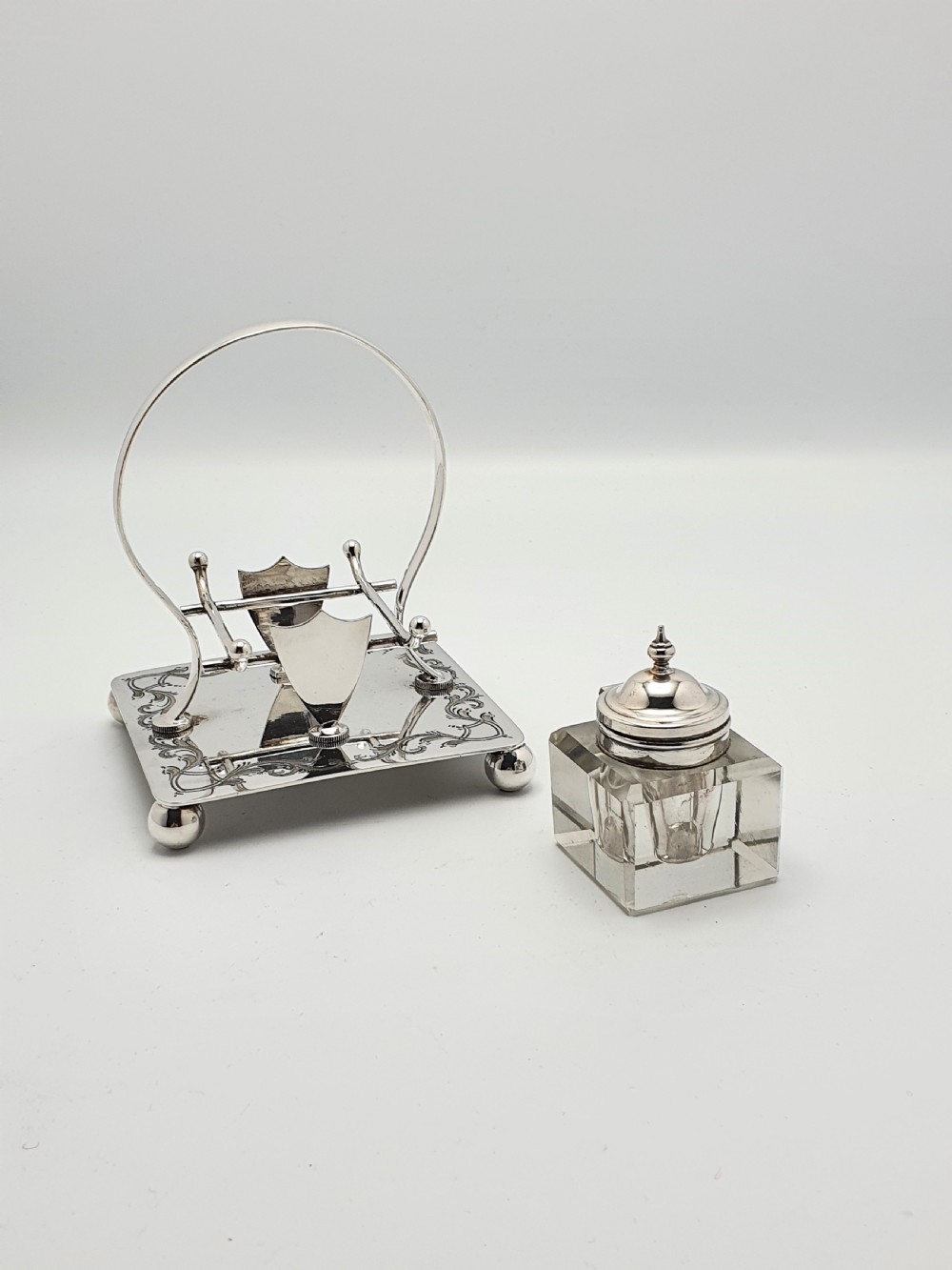 A Victorian silver-plated inkstand featuring an engraved square base on ball feet, a central clear cut-glass inkwell with a silver lid, an overhead hoop handle, and side pen rests. View V
