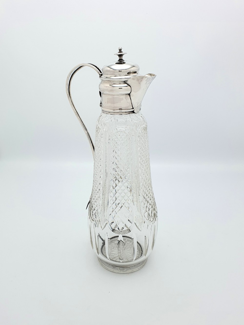 An antique Arts and Crafts period claret jug featuring a diamond-cut glass body with a silver-plated mount, hinged lid, and curved handle.