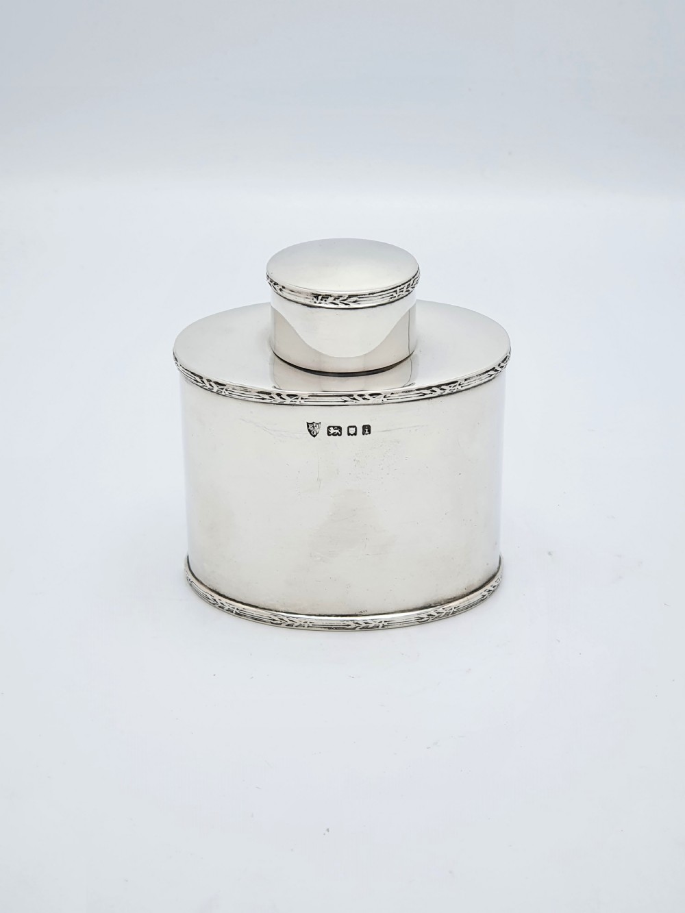 An oval sterling silver tea caddy with a cylindrical push-on cap, engraved leaf borders, and London hallmarks stamped on the front.