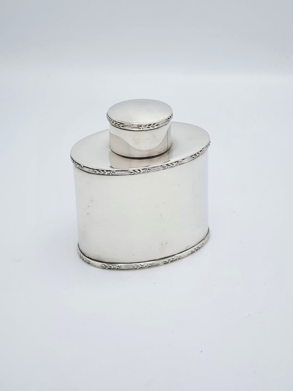 An oval sterling silver tea caddy with a cylindrical push-on cap, engraved leaf borders, and London hallmarks stamped on the front. View II