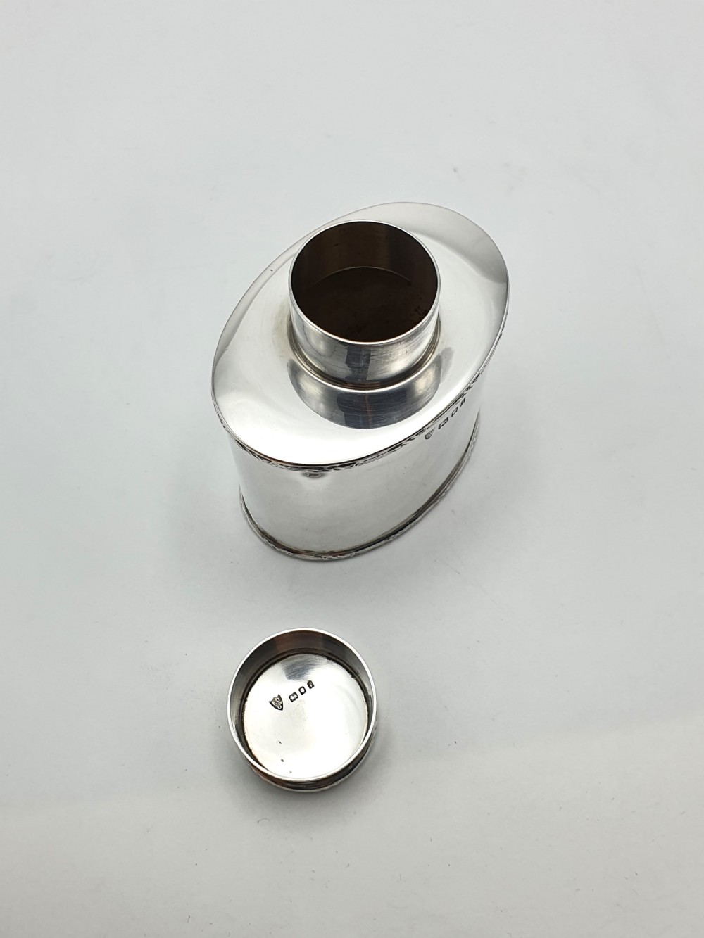 An oval sterling silver tea caddy with a cylindrical push-on cap, engraved leaf borders, and London hallmarks stamped on the front. View VI