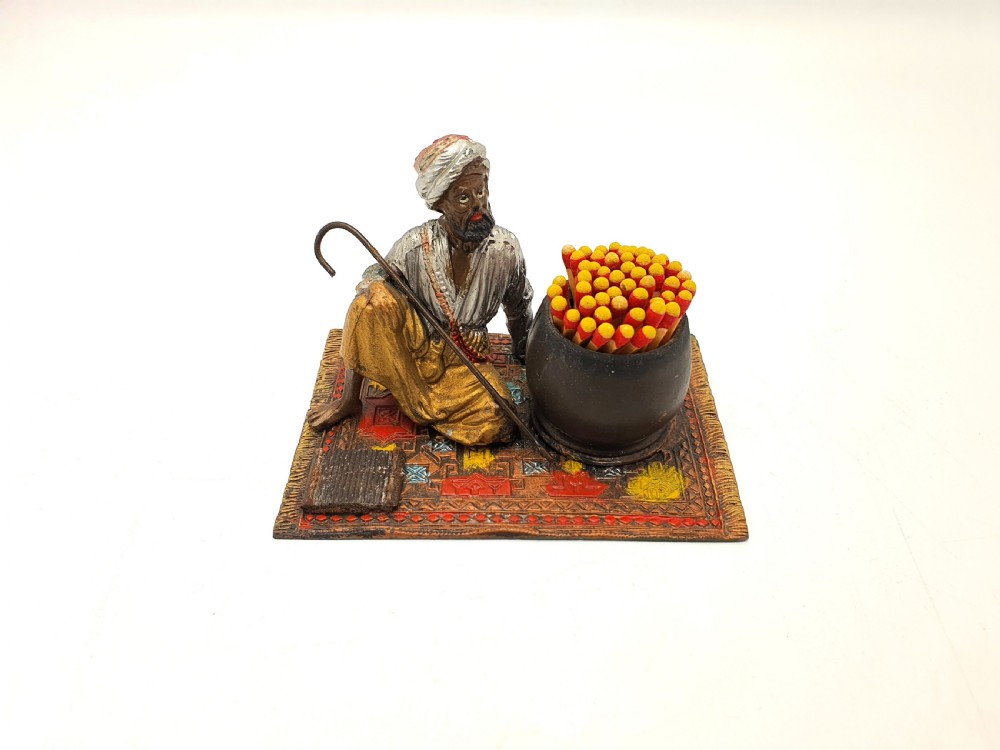 A cold-painted spelter figure of a seated Middle Eastern man with a staff on a patterned carpet base beside a match pot and striker pad.