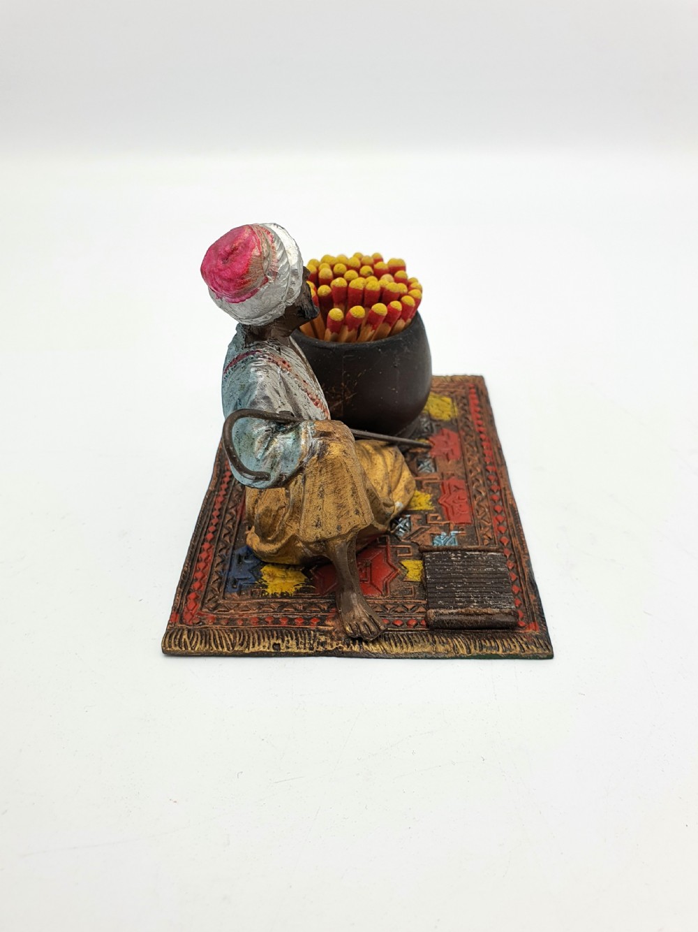 A cold-painted spelter figure of a seated Middle Eastern man with a staff on a patterned carpet base beside a match pot and striker pad. View II