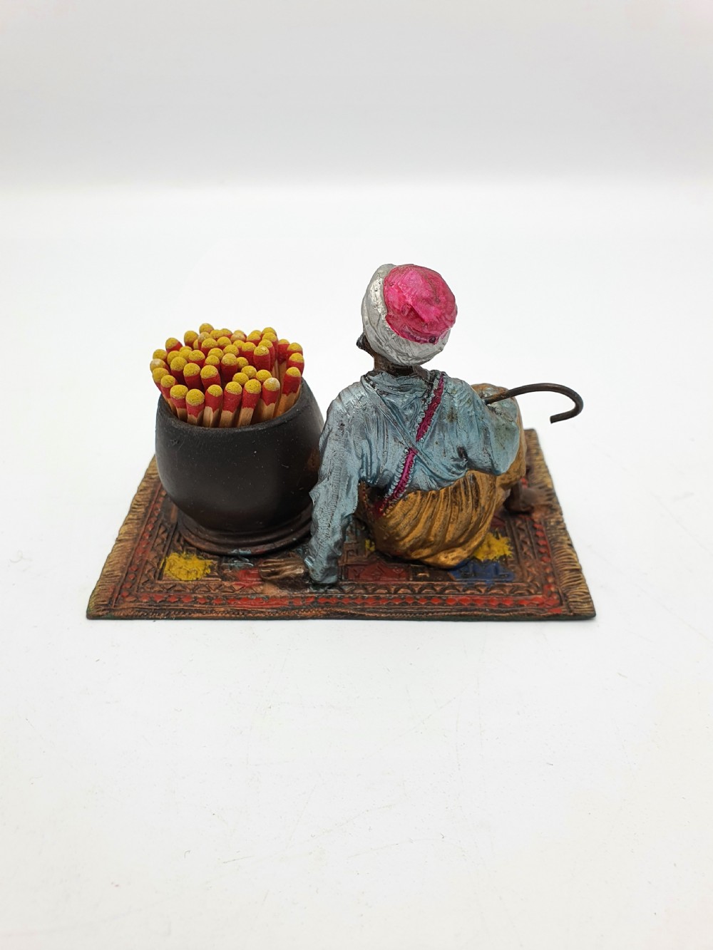 A cold-painted spelter figure of a seated Middle Eastern man with a staff on a patterned carpet base beside a match pot and striker pad. View III