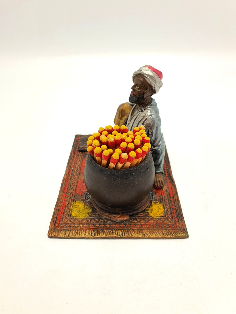A cold-painted spelter figure of a seated Middle Eastern man with a staff on a patterned carpet base beside a match pot and striker pad. View IV