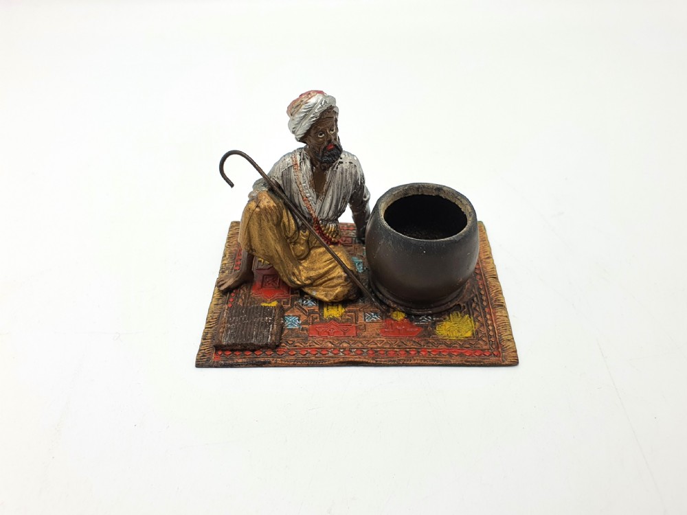 A cold-painted spelter figure of a seated Middle Eastern man with a staff on a patterned carpet base beside a match pot and striker pad. View V