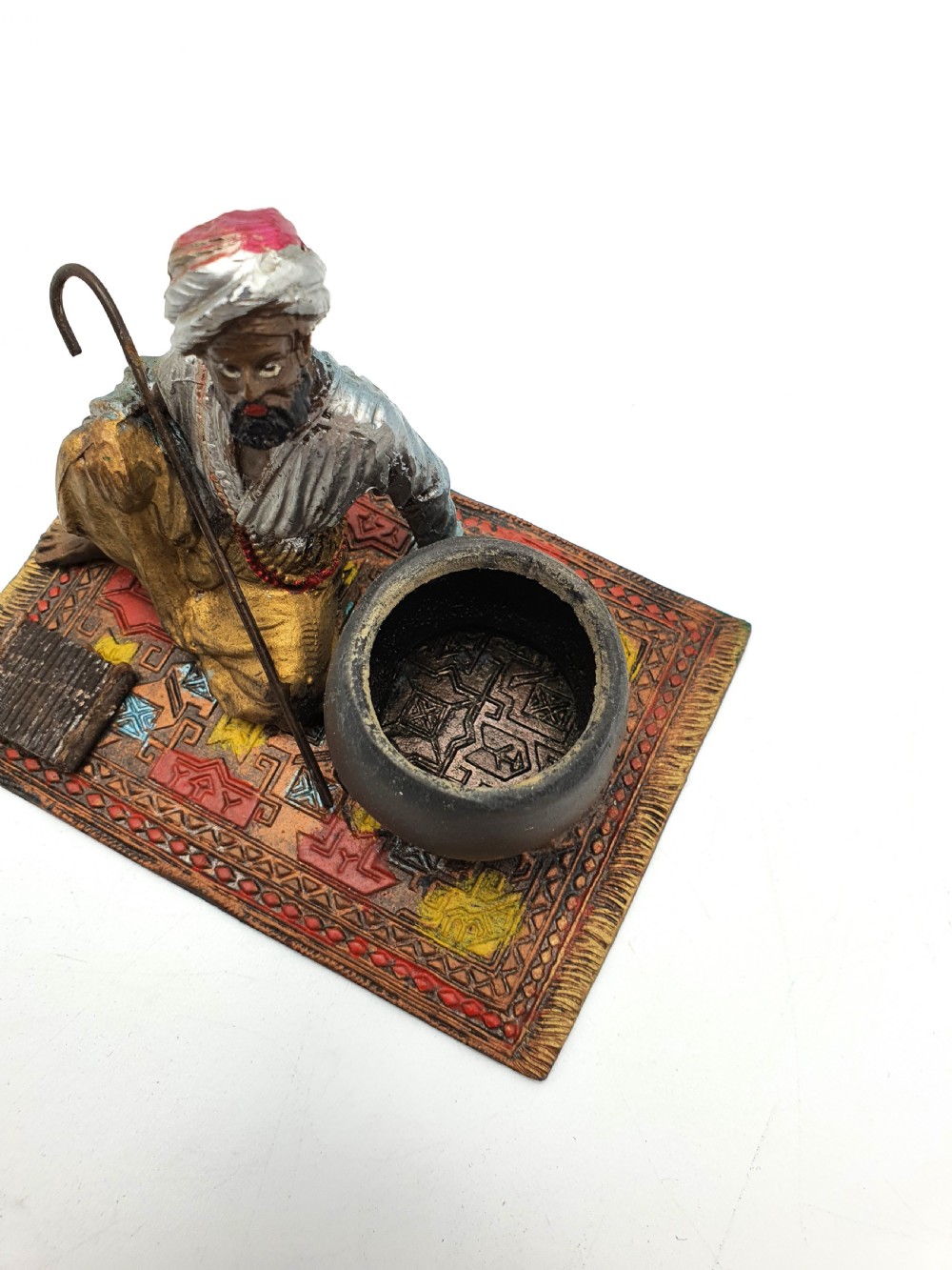 A cold-painted spelter figure of a seated Middle Eastern man with a staff on a patterned carpet base beside a match pot and striker pad. View VI