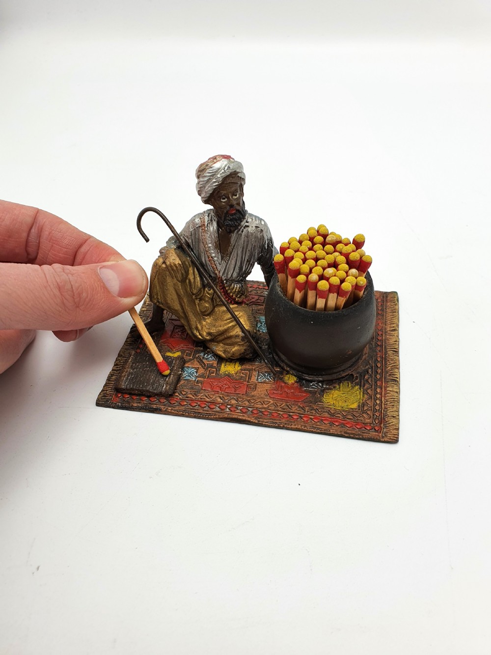 A cold-painted spelter figure of a seated Middle Eastern man with a staff on a patterned carpet base beside a match pot and striker pad. View VII