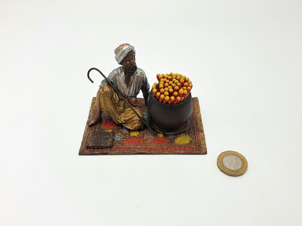 A cold-painted spelter figure of a seated Middle Eastern man with a staff on a patterned carpet base beside a match pot and striker pad. View VIII