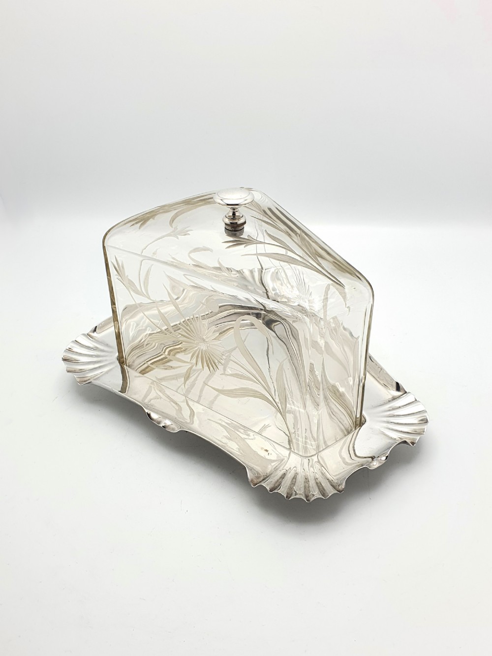 A wedge-shaped glass cheese dome with etched botanical details resting on a contoured silver-plated tray.