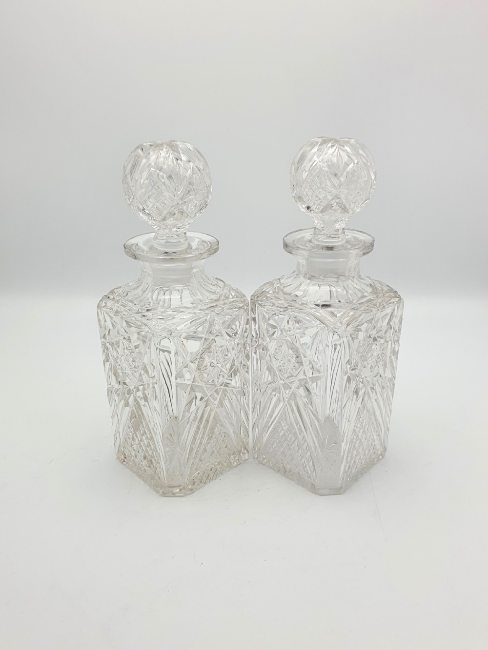 A pair of Edwardian clear cut glass spirit decanters with square bodies, geometric cut patterns, and matching spherical faceted stoppers.