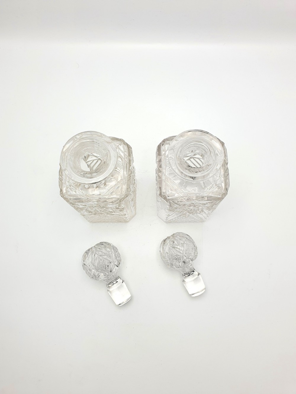 A pair of Edwardian clear cut glass spirit decanters with square bodies, geometric cut patterns, and matching spherical faceted stoppers. View V