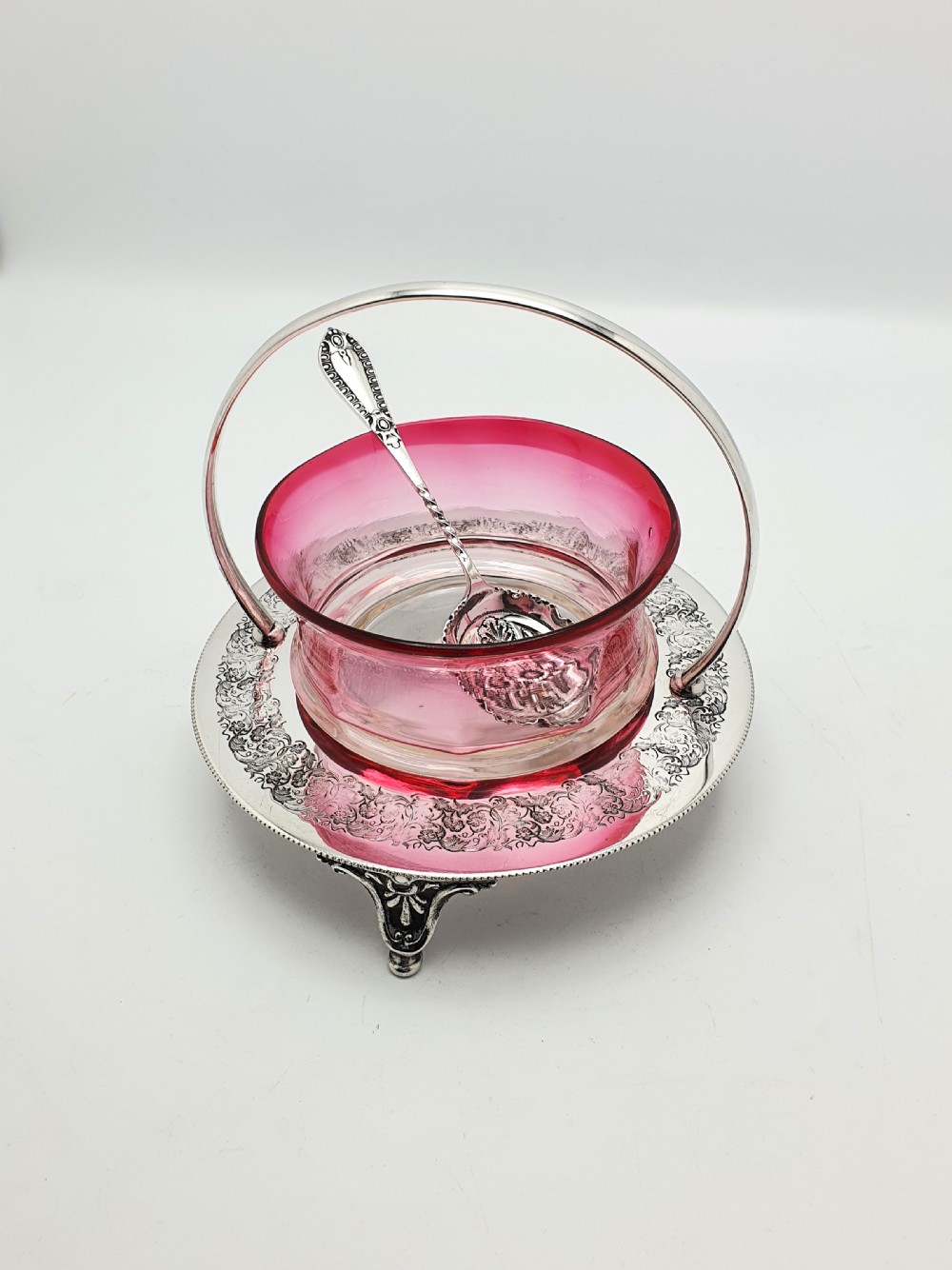 An Edwardian cranberry glass dish resting inside an engraved silver-plated stand with a swing handle and spoon.