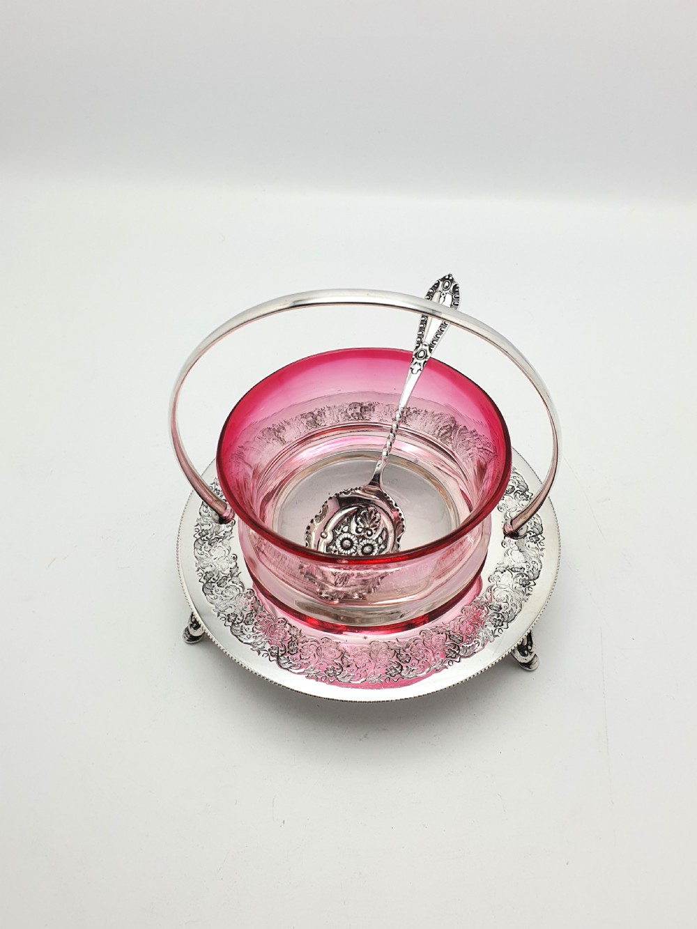 An Edwardian cranberry glass dish resting inside an engraved silver-plated stand with a swing handle and spoon. View II